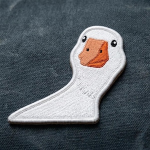 May include: Embroidered patch featuring a white goose with an orange beak and black eyes. The patch has a detailed design and is likely for application on clothing or accessories. The goose is facing left.