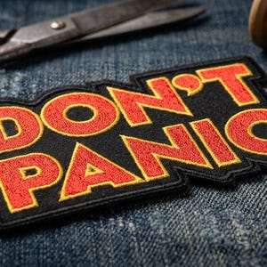 May include: Embroidered patch with the words "DON'T PANIC" in red letters with a yellow outline on a black background. The patch is on a denim surface, with scissors and a spool of gold thread in the background.