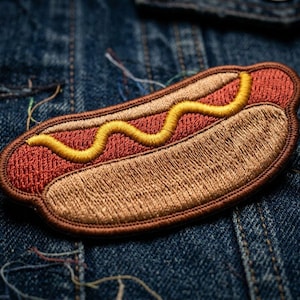 May include: Embroidered patch in the shape of a hot dog, featuring a red bun, a tan hot dog, and a yellow mustard squiggle. The patch is set against a blue denim background.