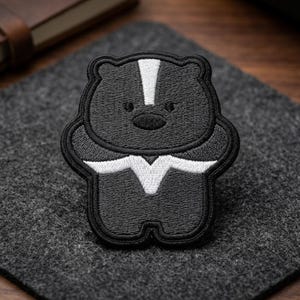 May include: Embroidered patch featuring a cartoon skunk design. The patch is black with white accents, including a stripe down the back and a chevron shape on the chest. The patch is on a gray felt surface.
