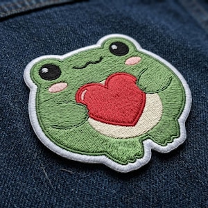 May include: Embroidered patch featuring a green frog holding a red heart. The frog has black eyes, pink cheeks, and a white outline. The patch is on a blue denim background.