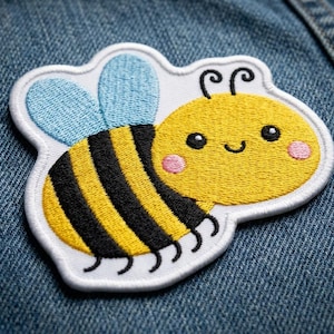 May include: Embroidered patch featuring a cartoon bee with a yellow body, black stripes, and blue wings. The bee has a smiling face with pink cheeks and is outlined in white. The patch is on a denim background.