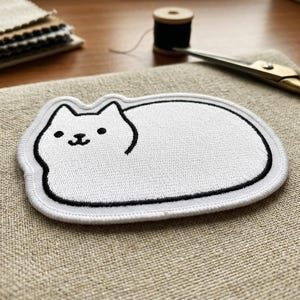 May include: White cat-shaped embroidered patch with a black outline and details. The patch is on a beige fabric surface, with sewing supplies like scissors and thread in the background. A cute, whimsical design.