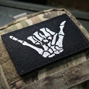 May include: A black rectangular patch featuring a white skeleton hand in the shaka gesture, sewn onto a camouflage fabric pouch. The patch is a unique accessory, perfect for adding a touch of personality to bags or clothing.