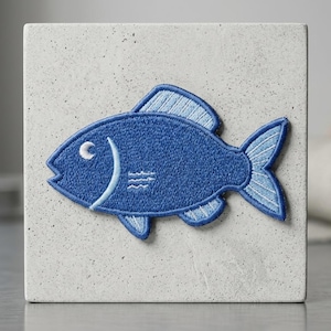 May include: A blue embroidered fish patch with light blue accents, displayed on a textured, light gray block. The fish has a curved body, a detailed tail, and a small eye. The patch is likely for decorative purposes.