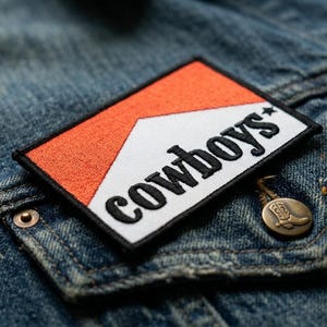 May include: Close-up of a denim jacket with a rectangular patch featuring the word "cowboys" in black lettering. The patch has an orange and white design. A small, bronze-colored pin with a cowboy boot is also visible.