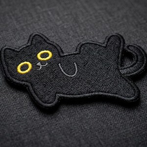 May include: Black embroidered cat patch with yellow eyes and a smiling face. The cat is lying on its back with its tail curled. The patch is on a dark gray textured background. A cute cat-themed accessory.
