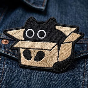 May include: Embroidered patch featuring a black cat peeking out of a cardboard box. The cat has white eyes and black paws. The patch is outlined in black and is sewn onto a denim jacket.