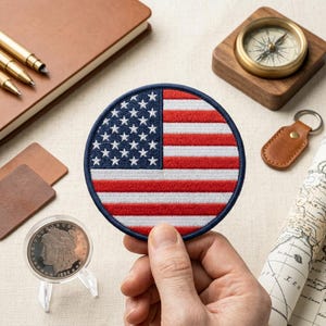 May include: A circular embroidered patch featuring the United States flag with a dark blue field of white stars on the left and horizontal red and white stripes on the right, finished with a dark blue stitched border around the entire edge.