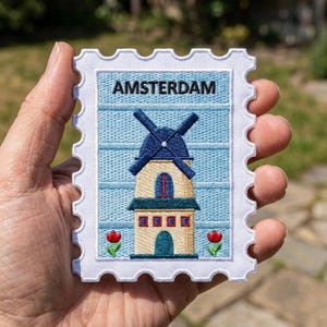 May include: A rectangular embroidered patch shaped like a postage stamp with a white border. The design features a light blue background with horizontal lines, a central windmill with a dark blue roof, and two small red tulips at the bottom. The text AMSTERDAM is embroidered in black at the top.