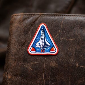 May include: Embroidered patch in a triangular shape featuring a space shuttle design. The patch has a white border with red accents, a blue background with white stars, and a white and blue shuttle.