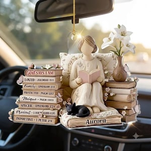 May include: A hanging car ornament featuring a faceless figure reading a book, seated on a stack of books with the name Aurora. The stack includes text: God Says I Am, Loved, Family, Protected, Unique, Special, Precious, and Important with Bible verses.
