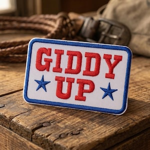 May include: A rectangular patch with the words "GIDDY UP" in red, outlined in white, and a blue border. Two blue stars flank the word "UP". The patch is on a wooden surface, with a rope and a bag in the background.