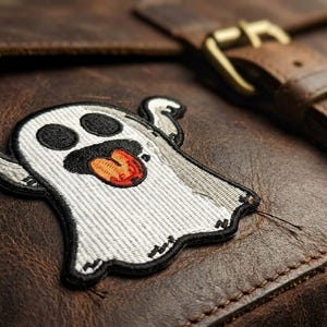 May include: A brown leather messenger bag with a stitched-on ghost patch. The ghost is white with black eyes and mouth, and a red tongue. The bag has a buckle closure and a brown leather strap.