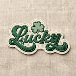 May include: Embroidered patch with the word "Lucky" in a green, stylized font. A green shamrock and Celtic knot are incorporated into the design. The patch has a cream-colored border and is set against a neutral fabric background.