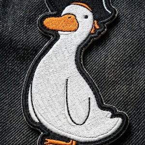 May include: Embroidered patch featuring a white duck wearing a black pirate hat with a skull and crossbones. The duck has an orange beak and feet, outlined in black. The patch is on a dark denim background.