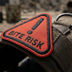 May include: A tactical dog harness with a red triangular patch. The patch has black trim and the words "BITE RISK" in white, with a black exclamation point. The harness is olive green and has a metal D-ring.