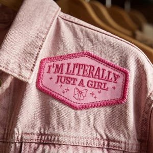 May include: A light pink denim jacket with a patch that reads "I'M LITERALLY JUST A GIRL" in pink lettering. The patch has a decorative bow and star design. The jacket is hanging on a wooden hanger.