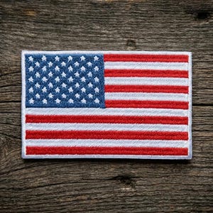 May include: Embroidered patch featuring the American flag. The flag has a blue field with white stars and red and white stripes. The patch is rectangular and has a white border. The background is a weathered wooden surface.