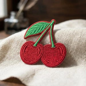 May include: Embroidered patch featuring two red cherries with green stems and a green leaf. The cherries have a detailed, textured design and a red border. The patch is placed on a beige fabric background.