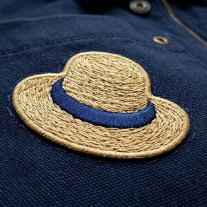 May include: Close-up of a navy blue fabric with an embroidered patch of a straw hat. The hat has a tan woven texture with a blue band. The patch is sewn onto the fabric with a matching blue thread.