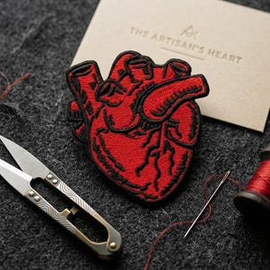 May include: A red embroidered heart patch with black outlining, a pair of small scissors, a needle, and a spool of red thread are arranged on a dark gray surface. A small card with the text "THE ARTISAN'S HEART" is also present.