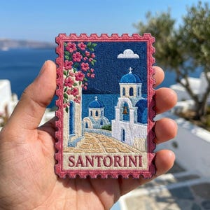 May include: A rectangular embroidered patch shaped like a postage stamp featuring a scene of Santorini with white buildings, blue domes, pink flowers, and a stone path. The word SANTORINI is stitched in red at the bottom with a pink scalloped border.
