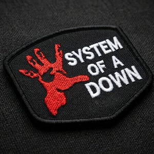 May include: Black patch with the text "SYSTEM OF A DOWN" in white, and a red hand graphic. The patch is embroidered with a black border. The background is a dark, textured fabric.