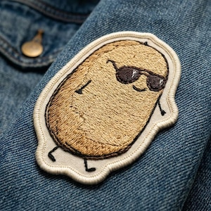 May include: Embroidered patch featuring a cartoon potato wearing sunglasses. The patch is tan with black details and a white border, sewn onto a denim jacket. The potato has arms and legs, giving it a playful appearance. The jacket is a medium wash blue.