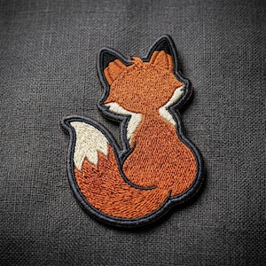 May include: Embroidered patch featuring a stylized fox in shades of orange, white, and black. The fox is depicted in a seated position with its tail curled. The patch has a black border and is likely designed for application on clothing or accessories.