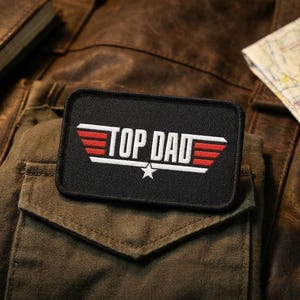 May include: A black patch with the text "TOP DAD" in white and red, featuring a star. The patch is on a green pocket, with a leather logbook and a map with a compass in the background. The items suggest a theme of aviation or adventure.