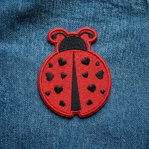 May include: A red ladybug patch with black heart-shaped spots and a black head. The embroidered patch is on a blue denim background. The ladybug's body is round, and the patch has a black border.