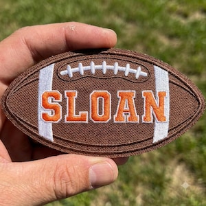 May include: An oval-shaped embroidered patch designed to look like a brown football with white laces and white vertical stripes. The name SLOAN is stitched in large, bold orange letters with a white outline across the center of the football patch.
