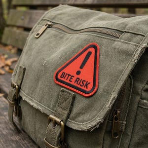 May include: An olive green canvas messenger bag with a red and black "BITE RISK" patch. The bag has multiple pockets, a zipper closure, and adjustable straps. The bag is sitting on a wooden bench.