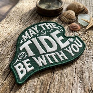 May include: Green embroidered patch with the text "MAY THE TIDE BE WITH YOU" in white. The patch is shaped like a stylized wave. A vintage compass, seashells, and a starfish are also visible.