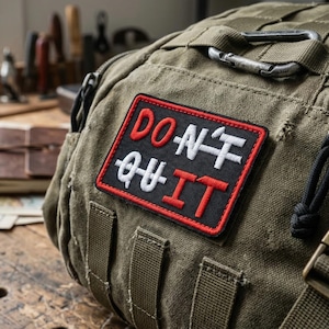 May include: Olive green duffel bag with a black, red, and white patch that reads "DON'T QUIT". The bag has multiple straps, a metal zipper, and a metal clasp. The bag is sitting on a wooden surface.