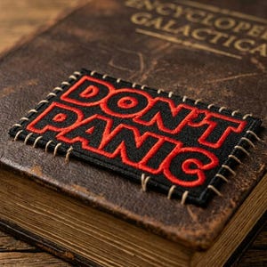May include: A vintage-style book with the title "ENCYCLOPEDIA GALACTICA" in gold lettering. A black and red embroidered patch that reads "DON'T PANIC" is sewn onto the cover. The book has a worn, brown leather cover.