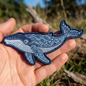 May include: A dark blue embroidered patch shaped like a humpback whale. The whale features intricate light blue thread detailing, including geometric patterns, wave-like lines, and small circular motifs across its body, fins, and tail fluke.