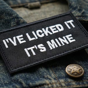 May include: A black rectangular patch with white embroidered text that reads "I'VE LICKED IT IT'S MINE" is affixed to a denim jacket. A bronze-colored button with a star design is visible below the patch.
