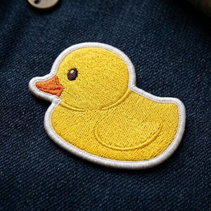 May include: Embroidered yellow duck patch with a white border, featuring an orange beak and a dark brown eye. The patch is sewn onto a dark blue denim fabric, showcasing a textured surface. A fun, whimsical accessory.