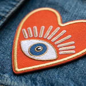 May include: An orange heart-shaped patch with an eye design, featuring a white eye with a blue iris and gold rays. The patch is embroidered with gold thread and is affixed to a denim jacket.