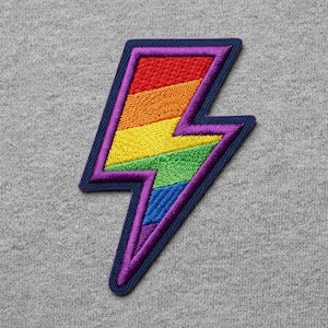 May include: Embroidered patch in the shape of a lightning bolt, featuring a rainbow design. The lightning bolt has a purple outline and a navy blue border. The rainbow colors include red, orange, yellow, green, blue, and indigo.