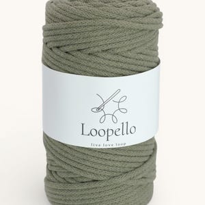 Op de afbeelding: Een spoel olijfgroen macramékoord. Het koord is in een cilindrische vorm om zichzelf gewikkeld. Een wit etiket wikkelt zich om het midden van de spoel met de merknaam "Loopello" en de slogan "live love loop".