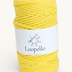 Op de afbeelding: Een spoel heldergeel garen met een wit etiket. Het etiket toont de merknaam "Loopello" en de slogan "live love loop" in zwart, samen met een naald- en draad-afbeelding. Het garen is strak opgerold.