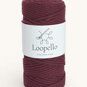Op de afbeelding: Een spoel bordeauxkleurig macramé-garen. Het witte etiket toont de merknaam "Loopello" en de slogan "live love loop" met een naald- en draad-afbeelding. Het garen is strak opgerold en toont zijn textuur.