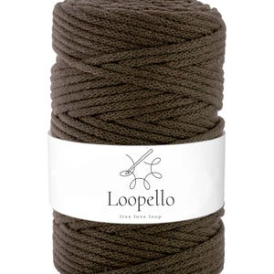 Può includere: Una bobina di cordoncino macramè marrone scuro. Il cordoncino è avvolto strettamente e avvolto con un'etichetta bianca che recita "Loopello" con un logo e il claim "live love loop". Il cordoncino sembra essere fatto di un materiale morbido e strutturato.