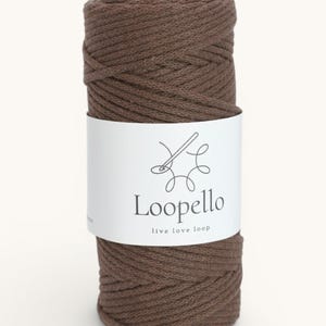 Può includere: Una bobina di cordoncino marrone per macramè. Il cordoncino è avvolto strettamente e avvolto con un'etichetta bianca. L'etichetta presenta un disegno a tratto di ago e filo, con la parola "Loopello" e la frase "live love loop".