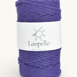 Op de afbeelding: Een rol paarse macramé koord. Een wit etiket wikkelt zich om het koord en toont de merknaam "Loopello" en de slogan "live love loop" in zwarte tekst. Het koord is een effen, levendige paarse kleur.
