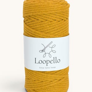 Può includere: Una bobina di cordoncino macramè giallo senape. Un'etichetta bianca avvolge il cordoncino, mostrando il marchio "Loopello" in un font moderno, con il claim "live love loop". Il cordoncino è ben avvolto, mostrando una superficie strutturata.