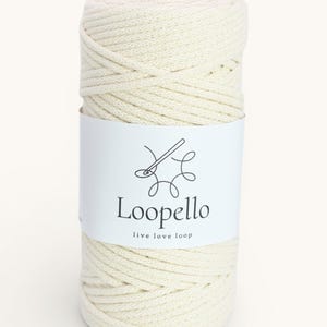 Può includere: Un rotolo di cordoncino macramè bianco sporco con un'etichetta bianca. L'etichetta mostra il marchio "Loopello" e il claim "live love loop", insieme a un logo di ago e filo. Il cordoncino è ben avvolto.